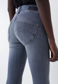 Jeans de mezclilla gris de cintura alta con corte ajustado, que cuentan con dos bolsillos traseros y detalles de costuras visibles. Textura suave y material resistente.