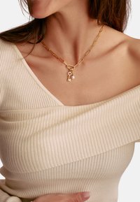 Goldkette mit großen Gliedern, einem herzförmigen Schlüsselanhänger und einem kleinen Perlenakzent. Tragen wird mit einem schulterfreien, gerippten Oberteil.