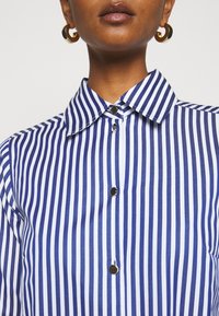 Camicia a righe blu e bianche con colletto, chiusura a bottoni e una texture del tessuto liscia. Decorata con bottoni metallici.