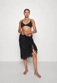 Top de biquíni preto e saia envelope combinando com detalhes em tassel, apresentando um tecido leve e um design até ao joelho. Modelo em pé descalça.