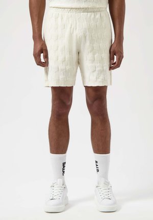 Pantaloni corti in spugna color crema con texture esagonale, vita elastica e tasche laterali, abbinati a sneakers bianche e calze con logo.