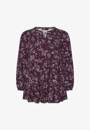 Fioletowa bluzka w kwiaty z długimi, bufiastymi rękawami, wycięciem w kształcie łezki z frędzlami oraz warstwowymi falbanami na dole. Wykonana z miękkiego materiału.