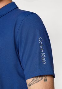 Kék póló, domború textúrájú anyagból, rövid ujjal, fehér "Calvin Klein" logóval a jobb vállon.