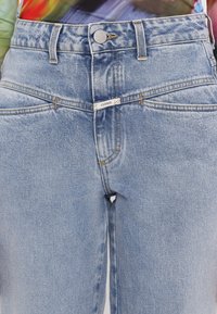 Heleda sinine kõrge vöökoht, teksapüksid, mis on valmistatud denimist, varustatud nupulehe ja esi-taskutega, nähtavate õmblusaktsentidega. Brändi silt on esitatud.