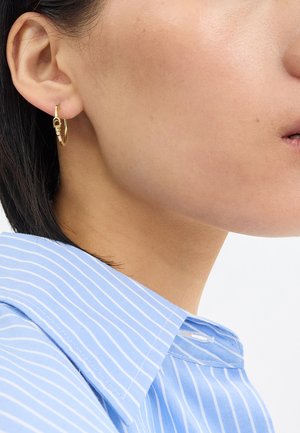 Orecchini a cerchio dorati con un piccolo gancio e superficie liscia e lucida, indossati da un modello con una camicia a righe blu e colletto.