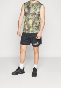 Haut de sport sans manches à motifs verts et marron, associé à un short noir. Des chaussures de sport noires avec des chaussettes blanches complètent la tenue.