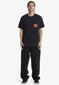 Svart bomullströja med korta ärmar, med ett rött logopatch på vänster bröst. Bärs med löst sittande svarta byxor och svarta sneakers.