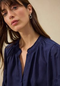 Camicia azzurra navy con colletto arricciato, maniche lunghe a sbuffo e dettagli di ricamo floreale sulle maniche. Tessuto con texture liscia.