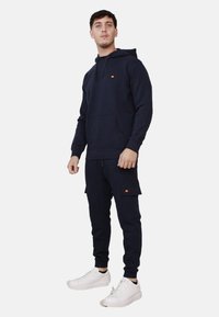 Felpa blu navy con tasca frontale e cappuccio, abbinata a pantaloni jogger coordinati con tasche cargo, indossata con sneakers bianche.