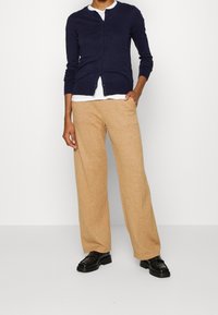 Merimiesneulospaita, jossa on viisi nappia, valkoisen paidan päällä. Beiget, leveälahkeiset housut pehmeästä materiaalista. Mustat nauhakenkäiset kengät täydentävät asun.