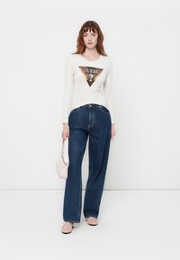 Camicetta a maniche lunghe color crema con un logo triangolare "GUESS" in colori metallici. Abbinata a jeans blu a vita alta e gamba larga, con accessori rosa chiaro.
