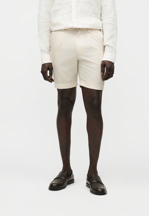 Mand iført beige opsmøgede shorts, hvid langærmet skjorte og mørkebrune læderloafers, stående mod en enkel hvid baggrund.