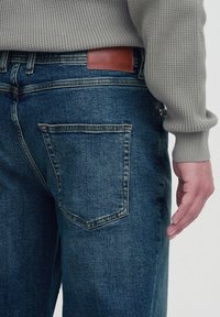 Jeanși din denim albastru cu o etichetă din piele la talie. Caracteristicile includ două buzunare spate și un mânecă de pulover tricotat gri texturat vizibil.