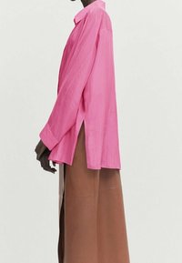 Roze, langdrogende shirt met een kraag, zijsplitten en een relaxte pasvorm. Gegdraag met een bruine, vloerlange rok van textuurmateriaal.