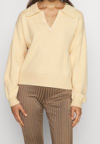 Ljusbeige stickad tröja med V-ringning, bred krage och sänkta axlar, i kombination med ribbade bruna byxor med vertikala ränder.