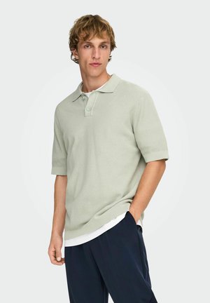 Mladý muž s rukou v kapse, oblečený v svetlozelenej textúrovanej krátkorukávovej polokošeli a tmavomodrých nohaviciach, stojaci pred jednoduchým pozadím.