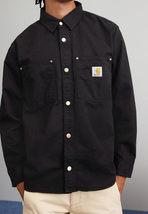 Mann trägt ein schwarzes Carhartt-Hemd mit Knöpfen und zwei Brusttaschen, kombiniert mit hellbeigen Hosen, vor einem einfachen grauen Hintergrund.