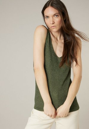 Femme aux longs cheveux bruns portant un haut en tricot vert sans manches et un pantalon blanc, les mains jointes devant, sur un fond neutre.