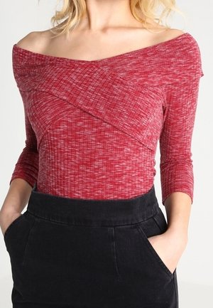 Rode off-shoulder top van een gestructureerde ribgebreide stof met driekwartmouwen en een diagonale crossover-ontwerp, gecombineerd met een zwarte rok.