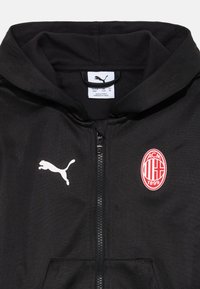 Camisola de capuz preta com fecho éclair, com o logótipo da Puma no lado esquerdo do peito e o emblema do clube de futebol AC Milan no lado direito do peito. Capuz visível na parte superior.