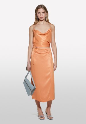 Robe en satin orange avec un décolleté en cowl et une taille cintrée, assortie à un sac à main bleu et des sandales à talons argentées. Texture lisse.