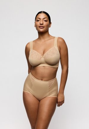 Vrouw met donker haar die een beige beha met kantaccenten en hoge tailleonderbroek modelleert tegen een effen lichte achtergrond.