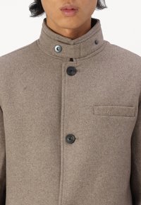 Abrigo de mezcla de lana marrón con un alto cuello, dos botones negros y un bolsillo en el pecho. Presenta un tejido texturizado y un diseño elegante.