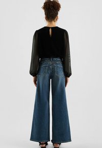 Zwarte fluwelen top met doorschijnende mouwen en een open rug met keyhole-detail. Lichtblauwe jeans met wijde pijpen en studs op de zakken en heupen.