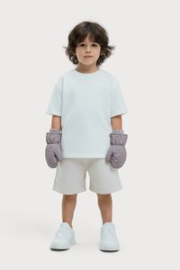 Dieťa s hnedými vlasmi, oblečené v bielom tričku, svetlých šortkách, bielych teniskách a veľkých šedých rukaviciach, stojí na jednofarebnom pozadí.