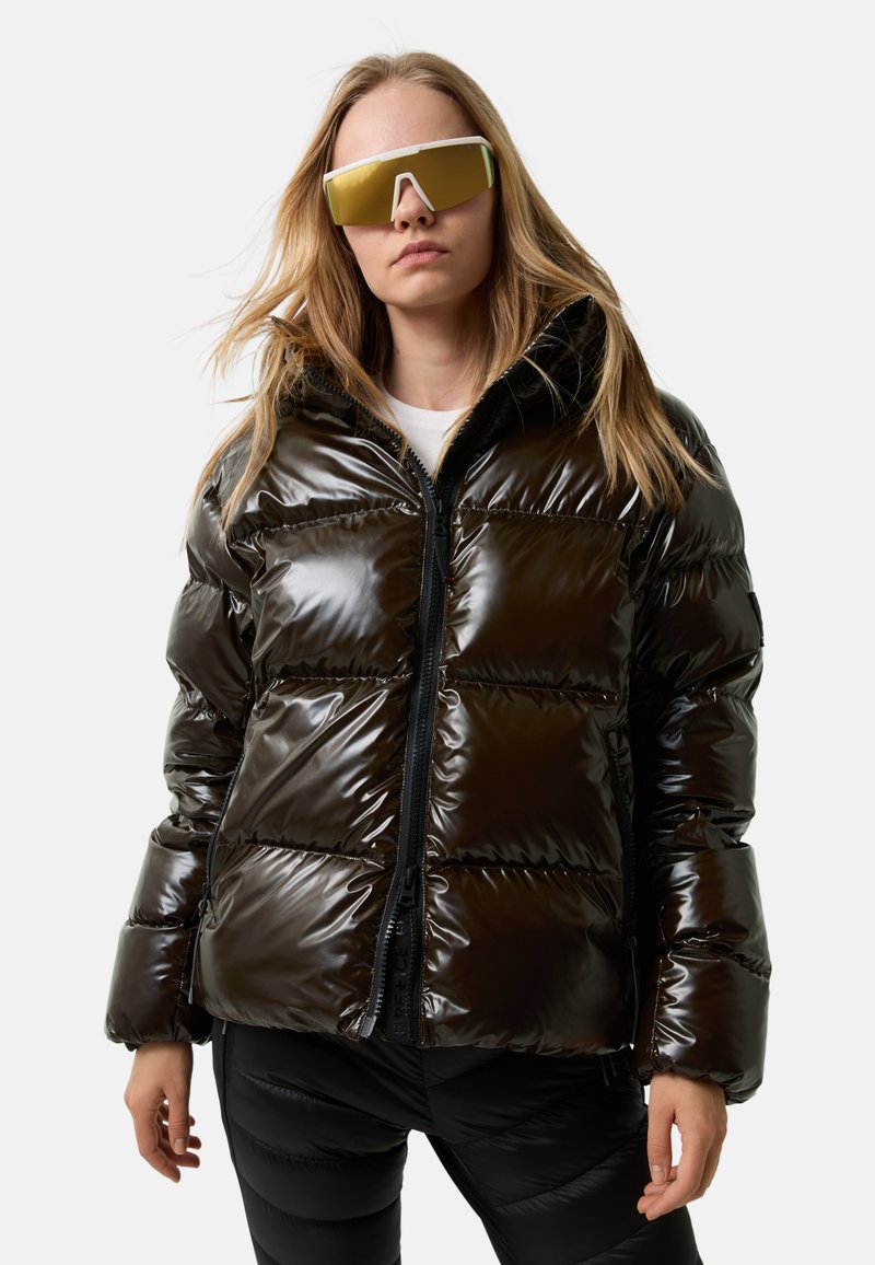 Kvinde iført en skinnende sort pufferjakke med høj krave og guldtonede oversized solbriller, stående foran en ensfarvet hvid baggrund.