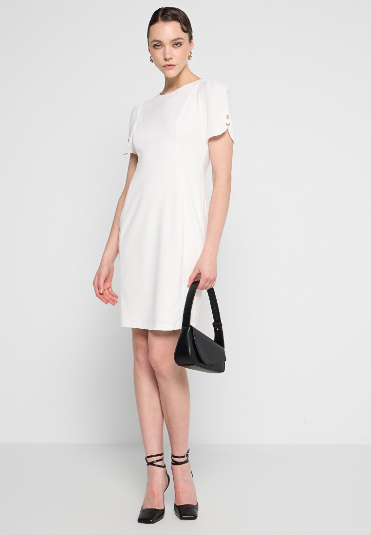 Krótka biała sukienka z krótkimi rękawami, ozdobiona złotymi guzikami, w połączeniu z czarną torebką i czarnymi sandałami na obcasie, na jednolitym tle.