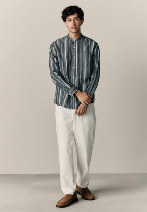 Young man with curly hair wearing a blue and white vertical striped shirt, white pants, and brown loafers, standing with hands clasped.