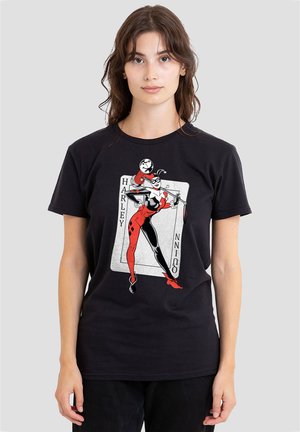 Young woman wearing black T-shirt showing red and black Harley Quinn comic character standing on a playing card background.