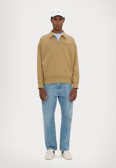 Beige quarter-zip sweatshirt with kangaroo pocket, worn over a light blue shirt, paired with light blue jeans and white sneakers. White cap on head.