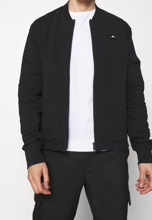 Homme portant une veste bomber noire zippée sur un t-shirt blanc avec un pantalon noir, debout les bras relâchés le long du corps.