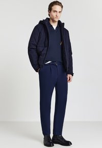 Tenue masculine comprenant une veste rembourrée bleu marine, un polo bleu foncé et un pantalon sur mesure. Des bottines noires brillantes viennent compléter le look.