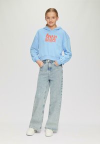 Niebieska, krótka bluza z kapturem z napisem "hey BENICE" w kolorze pomarańczowym, zestawiona z jasnoniebieskimi, szerokimi dżinsami i białymi sneakersami. Gładki materiał, luźny krój.