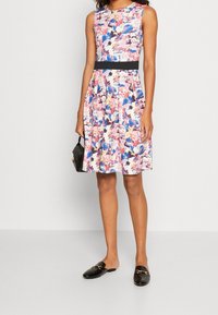 Robe sans manches à motifs floraux avec des nuances de rose, bleu et blanc ; cintrée à la taille avec une ceinture noire. Mocassins noirs plats et petit sac noir.