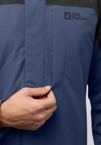 Veste imperméable bleue avec des accents noirs, dotée d'une poche zippée et d'une texture lisse. Logo "Jack Wolfskin" visible sur la poitrine.