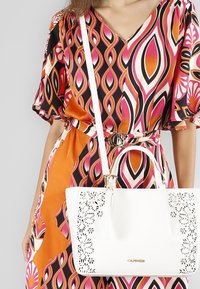 Borsa bianca con motivo floreale ritagliato e hardware metallico, abbinata a un vestito multicolore con motivi vorticosi in rosa, arancione e nero.
