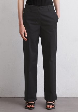 Vrouw draagt zwarte, recht gesneden broek met voorste knoop en zijzakken, gecombineerd met zwarte sandalen met open teen op een grijze vloer.