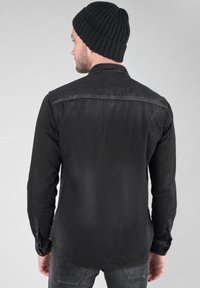 Camicia in denim nero con colletto a bottone, maniche lunghe e una texture leggermente scolorita, indossata con un berretto a coste nero.