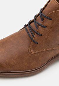Botín de cuero marrón con un acabado suave, puntera redonda y cordones oscuros, que presenta un diseño de costura a lo largo del borde superior y la suela.