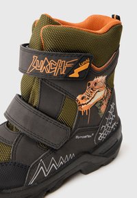 Botas infantiles en negro y verde oliva con detalles de malla, correas de velcro, gráfico de dinosaurio y un patrón en zigzag en la suela.