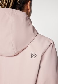 Veste à capuche rose clair avec une texture lisse, une capuche arrondie et un logo sombre à l'arrière. Design minimaliste avec des lignes épurées.