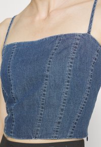 Farkkutoppi, jossa on ohuet olkaimet, pystysaumat, neliönmuotoinen pääntie ja tummansininen pesu.