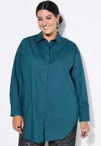 Teal langærmet skjorte med knapper, der har krave, to sideåbninger og manchetter. Stoffet virker glat med en let glans.