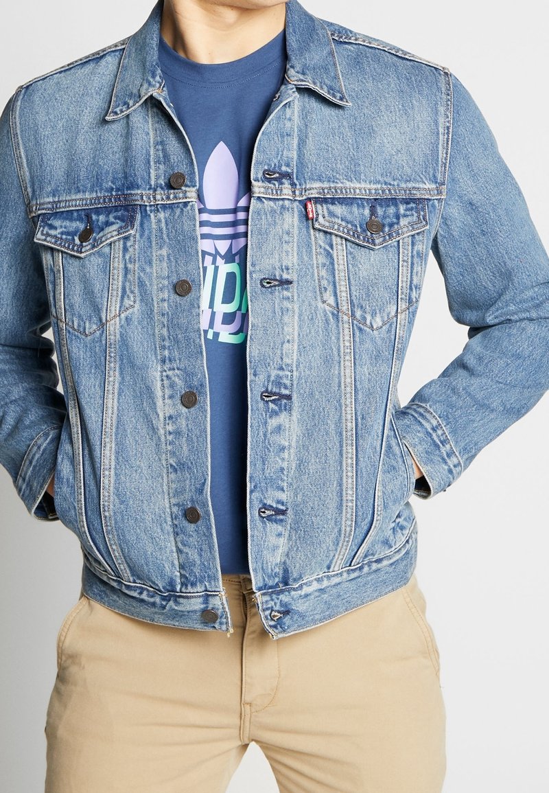 Chaqueta de mezclilla en azul claro, con cierre de botones, dos bolsillos en el pecho y un corte relajado. Combinada con una camiseta gráfica azul debajo.
