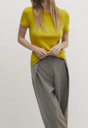 Woman wearing a short-sleeve yellow top with grey high-waisted wide-leg trousers, standing with hands adjusting the shirt hem.