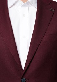 Burgundowy żakiet o strukturze z białą koszulą pod spodem, z czarną przypinką na klapie i okrągłym czarnym guzikiem.
